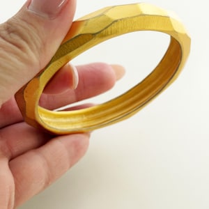 May include: A gold-colored bangle bracelet with a faceted, geometric exterior surface and a smooth, rounded interior. The metal features a brushed texture throughout, creating a matte finish on the angled edges of the circular band.