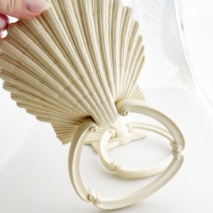 May include: Off-white seashell-shaped decorative handle with a fan-like design. The handle has a pair of curved, circular rings attached to the base. The handle is made of a solid material and is designed for use on a drawer or cabinet.