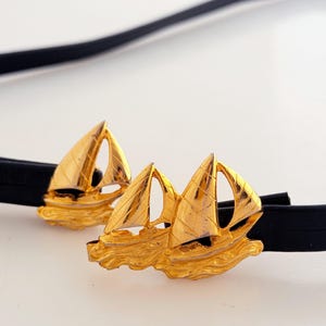 May include: A black belt featuring two gold-toned metal sailboat ornaments. Each ornament depicts a sailboat with textured sails and a stylized wave base. The belt strap is thin, smooth, and black, passing through the metal sailboat components.