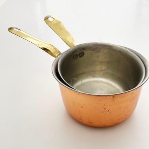 May include: Two small copper saucepans with brass handles. The pans are nested inside each other. The copper has a warm, reddish-brown color, and the brass handles have a golden hue. The pans are on a white surface.