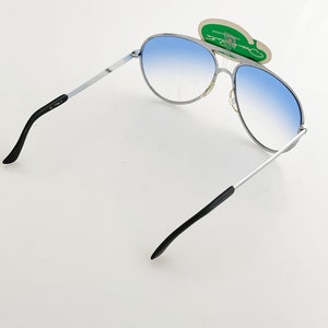 Op de afbeelding: Zonnebril in aviator-stijl met een zilverkleurig montuur en blauwe verlopende glazen. De zonnebril heeft zwarte pootjes en een label met de tekst "Oscar Ratti". De glazen lopen over van lichtblauw naar donkerder blauw.