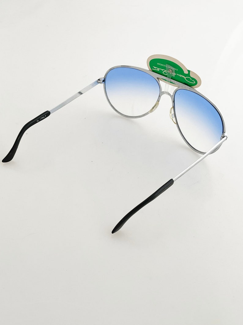 Op de afbeelding: Zonnebril in aviator-stijl met een zilverkleurig montuur en blauwe verlopende glazen. De zonnebril heeft zwarte pootjes en een label met de tekst "Oscar Ratti". De glazen lopen over van lichtblauw naar donkerder blauw.