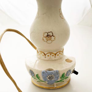 May include: A vintage ceramic lamp with a cream-colored base and a bulbous top. The lamp features gold trim and delicate floral designs in gold and blue. It has a brown electrical cord and a black on/off switch.