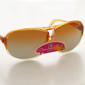 Può includere: Occhiali da sole aviator in plastica arancione con lenti marroni sfumate. Gli occhiali hanno un'etichetta attaccata che recita "Oscar de la Renta Fashion Eyewear" e "General Optics".