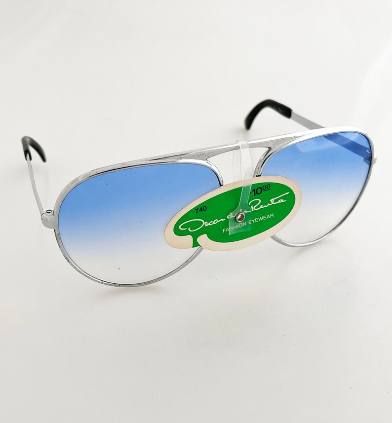 Op de afbeelding: Zilverkleurige pilotenbril met blauwe verlopende glazen. De zonnebril heeft zwarte oorschelpen en een groen-wit label met de tekst "Oscar de la Renta FASHION EYEWEAR".