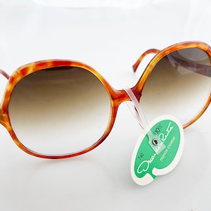 May include: Oversized, vintage sunglasses with a tortoiseshell pattern frame and gradient lenses. The sunglasses have a rounded shape and a tag that reads "Oscar de la Renta Fashion Eyewear".