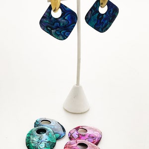 May include: A set of three pairs of earrings featuring iridescent abalone shell. The earrings are in the shapes of squares and teardrops. The earrings are displayed on a white stand.