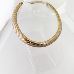 May include: A gold-toned omega chain necklace featuring a smooth, flexible, and rounded snake-like design. The necklace has a polished metallic finish with a continuous, segmented appearance that forms a circular shape around the neck display.