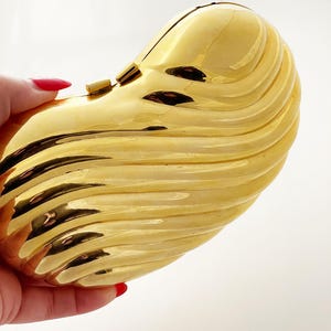 Può includere: Una pochette dorata con un design a coste a forma di onda. La pochette ha una forma arrotondata e una superficie lucida e riflettente. È dotata di una piccola chiusura a fermaglio. L'estetica generale è elegante e sofisticata.