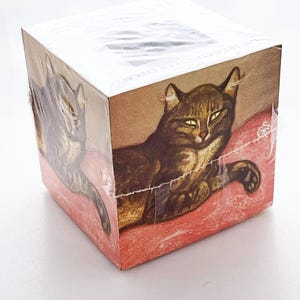 May include: A brown and white tabby cat with green eyes is lying on a red cushion. The cat is looking directly at the camera. The image is on a cube-shaped box with the text "The Artful Cat" on the side.