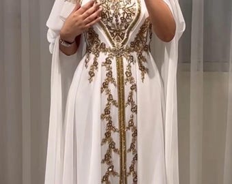 May include: A white formal gown with gold embroidery and embellishments. The dress features a V-neckline, a fitted bodice, and a flowing skirt. The gown has a matching head covering and cape-like sleeves. The text "Noori Boutique" is visible.