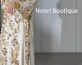 May include: A floor-length white kaftan featuring intricate gold and silver embroidery with sequins and beadwork. The garment has long, wide sleeves and a fitted waistline. The text Noori Boutique is visible on the right side of the image.