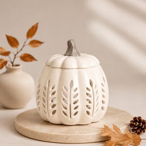 Può includere: Un barattolo in ceramica color crema a forma di zucca con coperchio rimovibile e stelo grigio. Il corpo del barattolo presenta intagli verticali con un motivo a foglie, che permettono alla luce di passare attraverso la struttura in ceramica.
