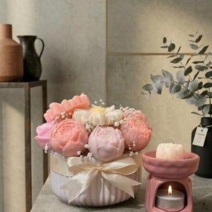 Puede incluir: Arreglo floral de flores de jabón rosas y blancas en un recipiente blanco con una cinta crema. Un quemador de aceite de cerámica rosa con una vela encendida y una flor de jabón está al lado. Jarrones decorativos en el fondo.
