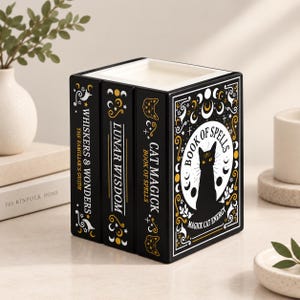 Może przedstawiać: Czarny ceramiczny pojemnik na świecę w kształcie stosu trzech książek. Z przodu znajduje się ilustracja kota z napisem Book of Spells oraz Magick Cat Energy. Grzbiety są oznaczone napisami Whiskers & Wonders, Lunar Wisdom oraz Cat Magick Book of Spells.