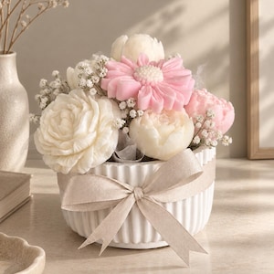 Pode incluir: Uma taça de cerâmica branca canelada preenchida com um arranjo floral de velas em forma de flor nas cores creme e rosa, intercaladas com pequenas flores brancas de gipsófila. Uma fita bege está atada em laço à volta do centro da taça.