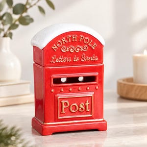 Puede incluir: Un buzón de cerámica rojo con una parte superior blanca cubierta de nieve. El frente presenta texto dorado en relieve que dice North Pole, Letters to Santa y Post. Tiene una ranura rectangular y una base decorativa con bordes dorados.