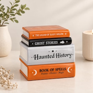 Op de afbeelding: Een stapel van vier decoratieve keramische boeken. Het bovenste boek is oranje met de tekst "THE LEGEND OF SLEEPY HOLLOW". Het tweede is zwart met "GHOST STORIES". Het derde is lichtgrijs met "Haunted History". Het onderste is oranje met "BOOK OF SPELLS".