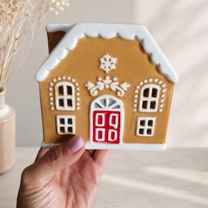 Può includere: Una statuetta a forma di casa di pan di zenzero in ceramica con esterno color marrone chiaro, dettagli di glassa bianca sul tetto e sulle finestre, una porta rossa centrale e un emblema a fiocco di neve bianco sopra la porta.