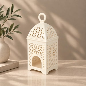 May include: A cream-colored ceramic lantern featuring an intricate laser-cut geometric pattern on all four sides and a domed top with a circular handle. The structure includes an arched opening at the base and a hollow interior for light.