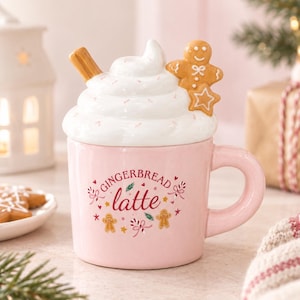 Può includere: Una tazza in ceramica rosa chiaro con la scritta GINGERBREAD latte in lettere rosse e rosa. Il coperchio ha la forma di panna montata bianca con una stecca di cannella marrone e un piccolo biscotto a forma di omino di pan di zenzero.