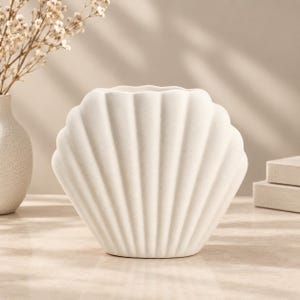 Könnte beinhalten: Eine matte, cremeweiße Keramikvase in Form einer Muschel mit vertikalen, geriffelten Rippen. Die Vase hat eine breite, geschwungene Silhouette und eine texturierte Oberfläche und dient als einzelnes Dekorationsobjekt mit einer Öffnung.