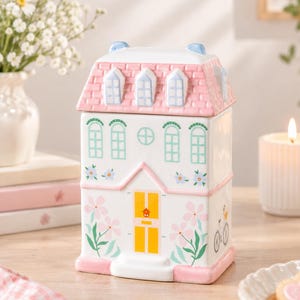 Può includere: Un contenitore in ceramica a forma di casa con coperchio rimovibile a tetto rosa. Il corpo bianco presenta finestre dipinte in azzurro, una porta gialla e motivi floreali rosa e verdi. Sul lato è visibile una piccola illustrazione di bicicletta.
