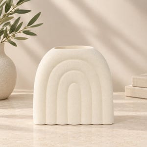 Puede incluir: Un jarrón de cerámica de color crema con un acabado moteado y una forma de arco distintiva. La superficie frontal presenta tres curvas concéntricas en relieve similares a un arcoíris, creando una apariencia texturizada con una abertura superior ancha.