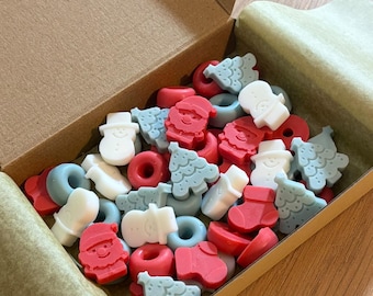 Può includere: Una scatola di cartone riempita con piccole cere profumate a tema festivo in varie forme, tra cui volti di Babbo Natale rossi, pupazzi di neve bianchi, alberi di Natale azzurri, calze rosse e anelli azzurri, disposti all'interno del contenitore.