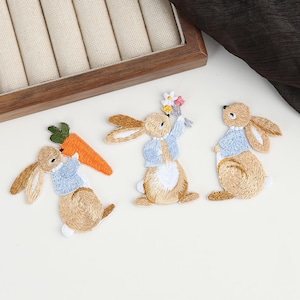 May include: Three embroidered bunny patches. Each bunny is light brown with tan ears and a light blue jacket. One bunny holds a carrot, another holds flowers, and the third stands upright. The patches are likely for clothing or craft projects.