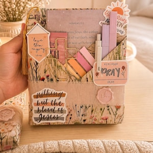 May include: A fabric book cover with floral and grass patterns, holding a book and various stationery items. The cover has pockets and decorative stickers with phrases like "love them anyway" and "nothing but the blood of Jesus."