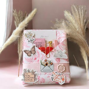 May include: A gift box wrapped in pink floral paper, adorned with stickers including a butterfly, flowers, and the text "believe." A red heart-shaped cross and the text "John 14:6-7" are visible.