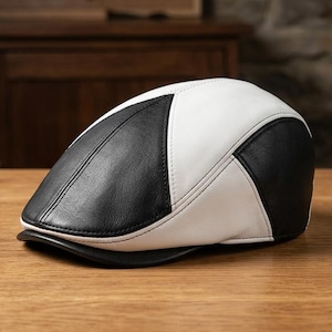 May include: A flat cap constructed from alternating panels of smooth black and white leather. The cap features a rounded crown, a short stiffened brim at the front, and visible stitched seams connecting the contrasting monochrome leather sections.