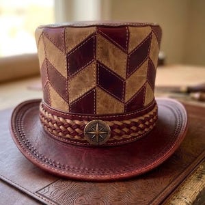 May include: A handcrafted leather top hat with a burgundy and tan chevron pattern. The hat features a braided band with a compass rose emblem and a wide brim. The hat is displayed on a leather surface.