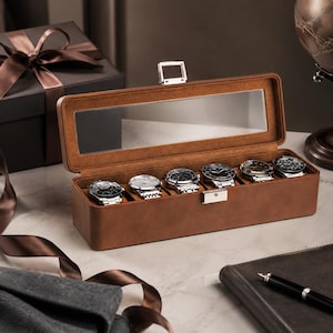 May include: A rectangular brown watch storage box with a clear glass lid and a silver metal latch. The interior features six individual compartments, each containing a wristwatch with a metallic band. The box has a smooth, matte finish.
