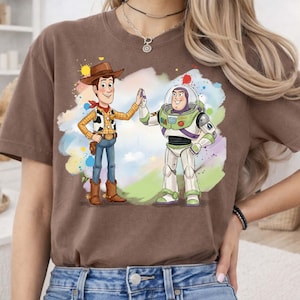 May include: A brown short-sleeve t-shirt featuring a graphic print of Woody and Buzz Lightyear high-fiving. The characters are set against a colorful watercolor-style splash background with shades of blue, green, yellow, and red accents.