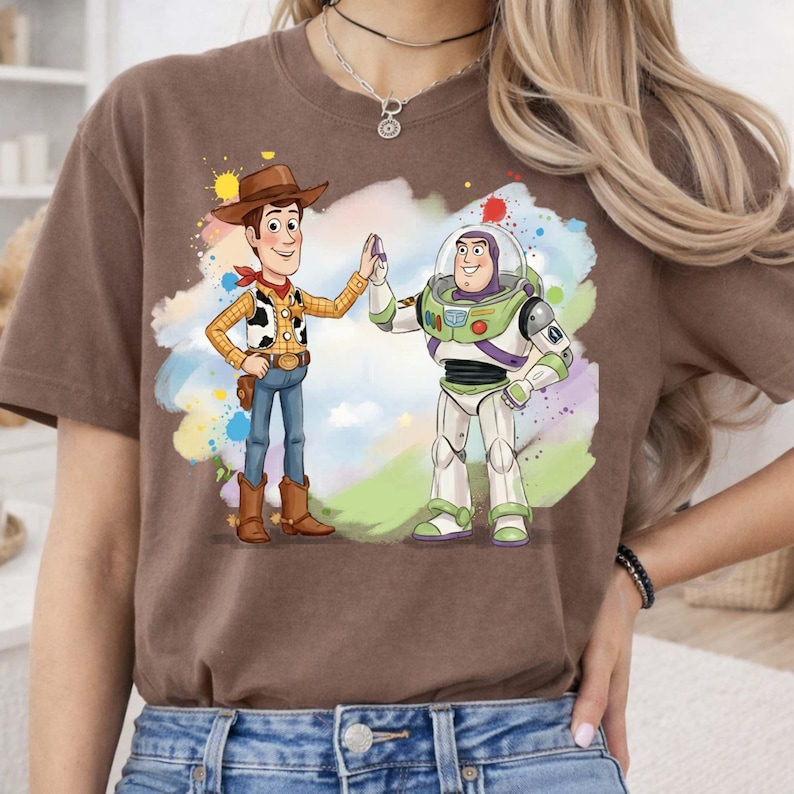 May include: A brown short-sleeve t-shirt featuring a graphic print of Woody and Buzz Lightyear high-fiving. The characters are set against a colorful watercolor-style splash background with shades of blue, green, yellow, and red accents.