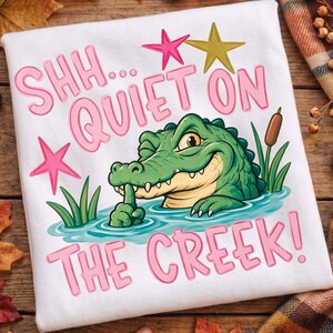 Może przedstawiać: Biały t-shirt z grafiką zielonego aligatora w niebieskiej wodzie z zieloną trzciną. Napis SHH... QUIET ON THE CREEK! wykonano różowymi literami. Nad aligatorem, który trzyma palec przy ustach, znajduje się różowa i złota gwiazda.