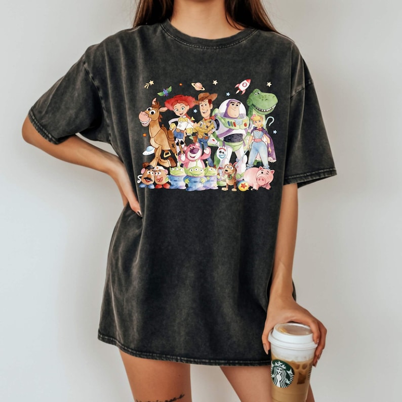 May include: Dark gray t-shirt featuring a colorful graphic of characters from the animated movie Toy Story. The design includes Woody, Buzz Lightyear, and other beloved characters. The shirt has a vintage, washed look.