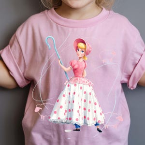 May include: A light pink t-shirt featuring a graphic print of a character with blonde hair wearing a pink bonnet, a pink bodice, and a white skirt with pink polka dots. The character holds a light blue shepherd's crook, surrounded by faint outlines of sheep.
