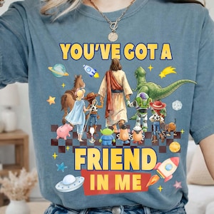 May include: Blue t-shirt with the text "YOU'VE GOT A FRIEND IN ME" in yellow. The graphic features cartoon characters, including Jesus, a dinosaur, and space-themed elements. A rocket ship and a UFO are also included.