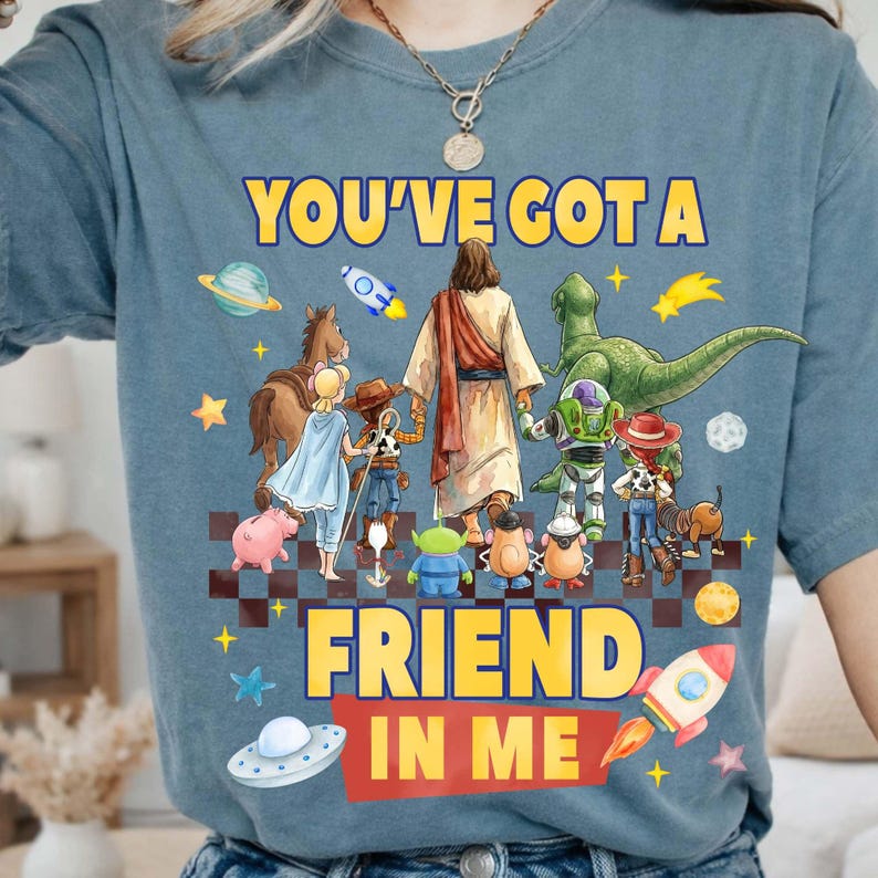May include: Blue t-shirt with the text "YOU'VE GOT A FRIEND IN ME" in yellow. The graphic features cartoon characters, including Jesus, a dinosaur, and space-themed elements. A rocket ship and a UFO are also included.