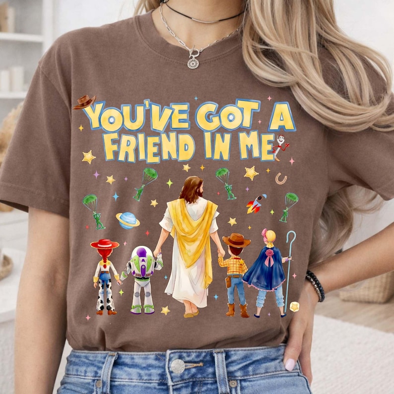 May include: Brown t-shirt with the text "YOU'VE GOT A FRIEND IN ME" in yellow. The graphic features cartoon characters and a figure in white robes holding hands with the characters. Stars and other small graphics are scattered around the text.