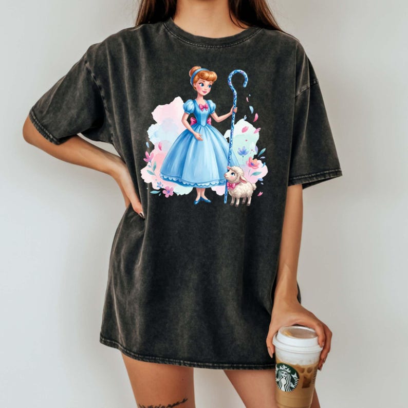 May include: A charcoal grey acid-wash t-shirt featuring a graphic print of a character in a blue dress holding a blue shepherd's crook, standing next to a small white lamb. The design includes colorful watercolor-style splashes in pink and blue.