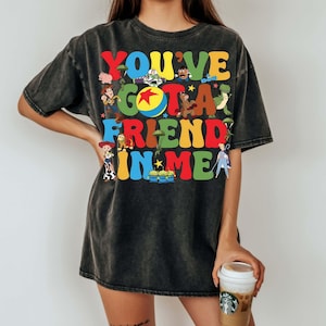 May include: Dark gray t-shirt featuring the phrase "YOU'VE GOT A FRIEND IN ME" in colorful, cartoon-style lettering. The design includes various character illustrations. The shirt is a relaxed fit.