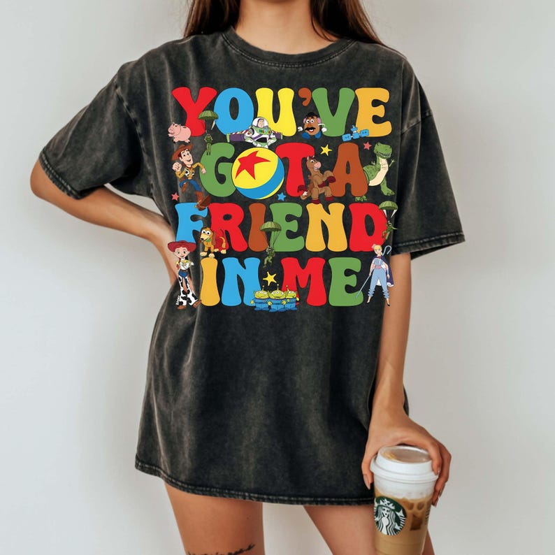 May include: Dark gray t-shirt featuring the phrase "YOU'VE GOT A FRIEND IN ME" in colorful, cartoon-style lettering. The design includes various character illustrations. The shirt is a relaxed fit.