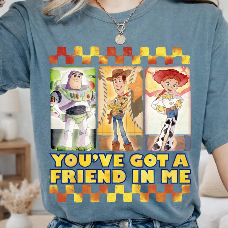 May include: A light blue t-shirt featuring three characters from the Toy Story movie: Buzz Lightyear, Woody, and Jessie. The text "YOU'VE GOT A FRIEND IN ME" is printed in yellow and blue. The design is framed by a red and yellow checkered pattern.