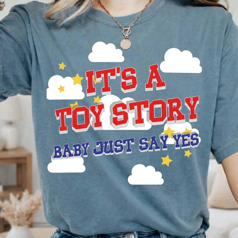 以下が含まれることがあります： 赤と白のテキスト「IT'S A TOY STORY BABY JUST SAY YES」がプリントされた青い半袖Tシャツ。デザインには白い雲のグラフィックと黄色い星のアクセントが前面に散りばめられており、レイヤー状の視覚効果を生み出しています。