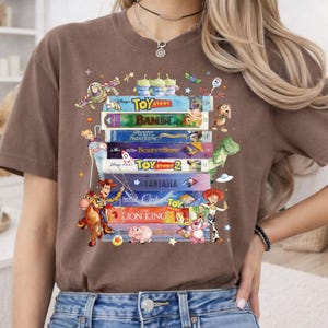 May include: A brown t-shirt featuring a stack of classic Disney VHS tapes, including "Toy Story," "Bambi," and "The Lion King." The design incorporates cartoon characters like Buzz Lightyear, Woody, and Rex, with a whimsical, colorful aesthetic.