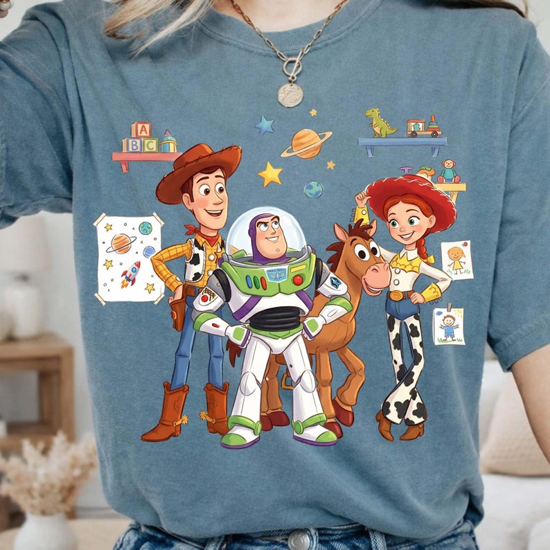 May include: A blue t-shirt featuring a graphic print of Woody, Buzz Lightyear, Bullseye, and Jessie from Toy Story. The design includes colorful illustrations of planets, stars, and toy shelves with blocks, a dinosaur, and children's drawings.